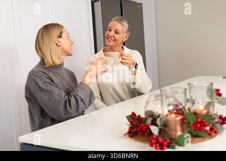 Mother and daughter clinking beige mugs while talking at home kitchen island near candle display Stock Photo