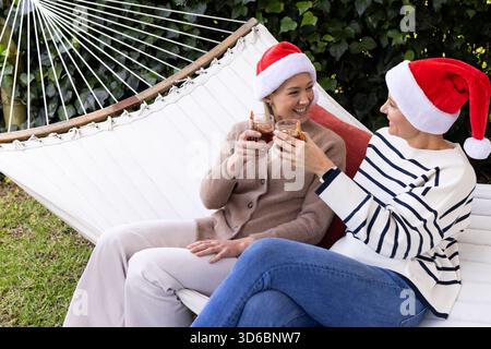 Female friends wearing Santa hats sitting on hammock clinking glasses of dark cola in backyard Stock Photo