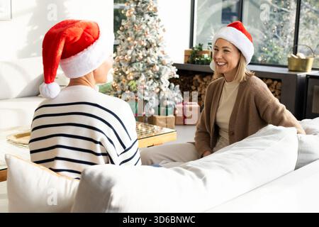 Mother and daughter chatting on couch in living room with Santa hats, decorated Christmas tree Stock Photo