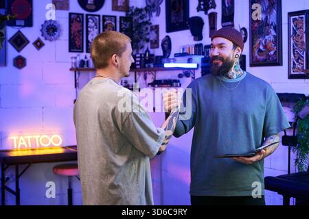 Caucasian young adult man with tattoos shaking hands with tattooed Caucasian middle aged man wearing beanie in tattoo studio, both smiling and making eye contact Stock Photo