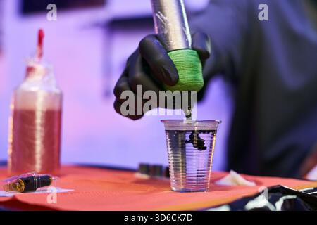 Black gloved hand of young adult Caucasian man holding tattoo machine and dipping needle into plastic cup with black ink, preparing for tattooing session in professional studio Stock Photo