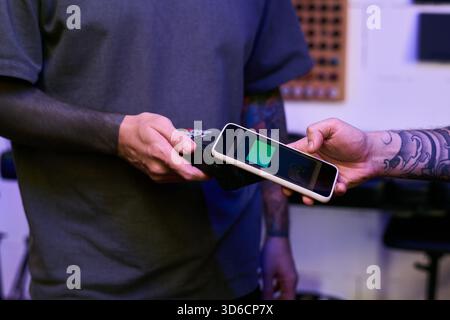 Caucasian young adult man with tattooed arms holding payment terminal while another Caucasian young adult man with tattoos making contactless payment using smartphone Stock Photo