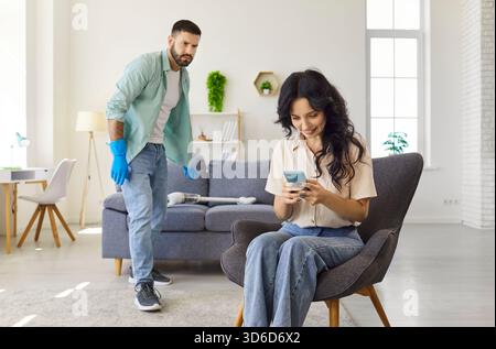 Young couple at home, man vacuuming while woman uses smartphone in living room Stock Photo