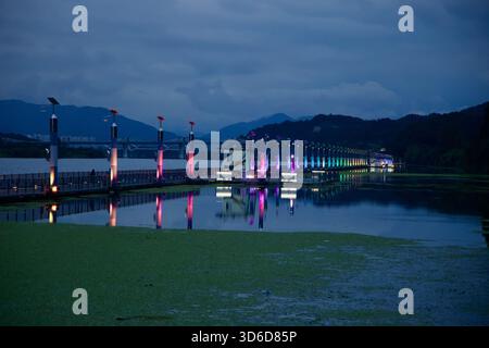 Inside the Rainbow Bridge approach, rows of paper lanterns in red, green, blue, and yellow hang over a winding brick path as the evening sky darkens a Stock Photo