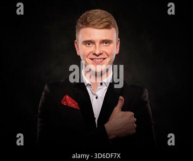 A smiling young redheaded man wearing a formal black suit and giving a thumbs up gesture of approval against a dark background. Stock Photo