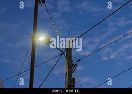 An illuminated LED streetlight attached to a concrete utility pole, shining brightly against a deep blue evening sky with soft clouds and many wires. Stock Photo