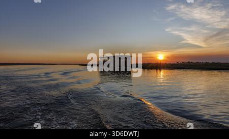 Aerial view of the sun setting on the Chobe River, casting a golden glow over the tranquil waters and a houseboat creating ripples as it glides, Kasan Stock Photo