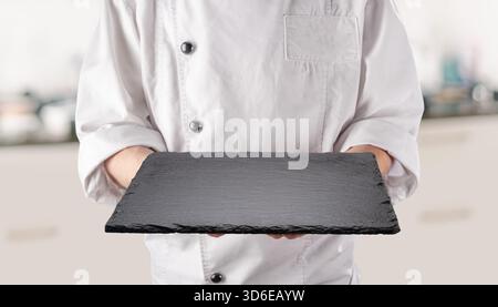 Chef in white uniform is holding an empty black slate serving plate. Black background. Stock Photo