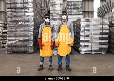 Two industrial workers wearing protective gear including yellow aprons, face masks, safety goggles, and gloves pose confidently in a warehouse setting Stock Photo