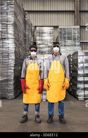 Two industrial workers wearing protective gear including yellow aprons, face masks, safety goggles, and gloves pose confidently in a warehouse setting Stock Photo