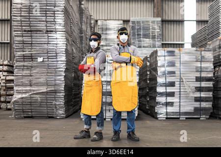 Two industrial workers wearing protective gear including yellow aprons, face masks, safety goggles, and gloves pose confidently in a warehouse setting Stock Photo