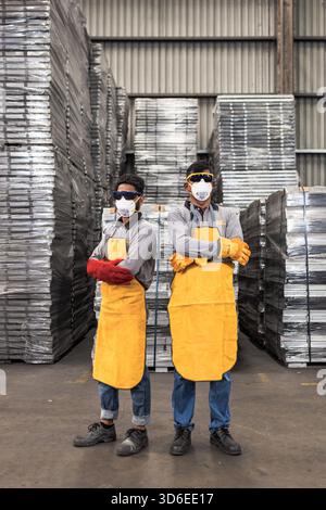 Two industrial workers wearing protective gear including face masks, safety goggles, long-sleeved shirts, yellow aprons, and heavy-duty gloves pose co Stock Photo