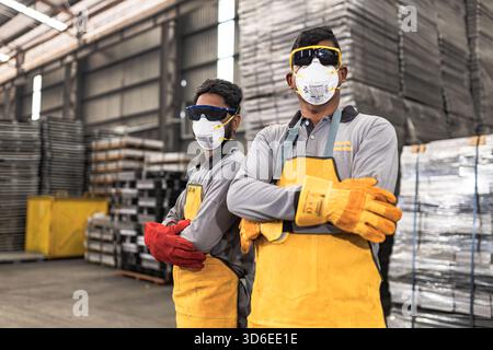 Two industrial workers wearing protective gear including face masks, safety goggles, long-sleeved shirts, yellow aprons, and heavy-duty gloves pose co Stock Photo
