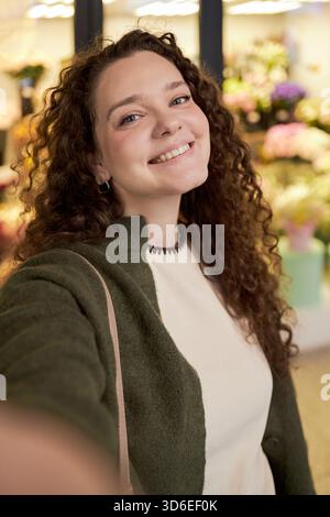 Portrait of young adult Caucasian woman smiling and taking selfie in flower shop, curly hair framing face, colorful floral arrangements visible in background, cheerful expression Stock Photo