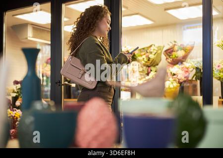 Young adult Caucasian woman standing in front of flower shop window holding smartphone, looking at screen, surrounded by colorful floral arrangements and decorative vases Stock Photo