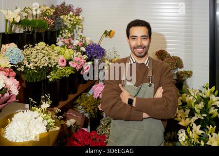Young adult Black man standing with arms crossed in front of colorful flower arrangements in florist shop, smiling confidently, surrounded by various fresh blooming flowers Stock Photo