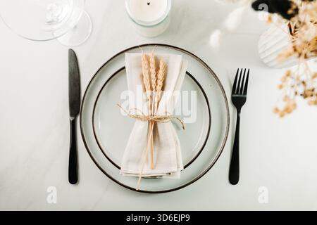 Elegant minimal table setting with ceramic plates, black cutlery and a linen napkin decorated with wheat stems, styled in a soft natural light. Stock Photo