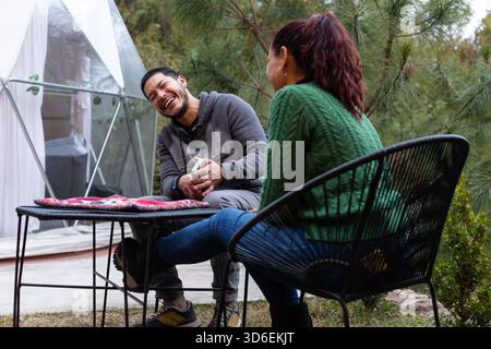 Two people are enjoying a lively conversation outdoors, seated in a cozy garden area next to a glamping dome. They appear relaxed and cheerful, surrou Stock Photo