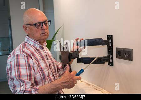 An elderly man is focused on DIY installation of a TV wall mount, using a screwdriver and examining the bracket carefully. Stock Photo