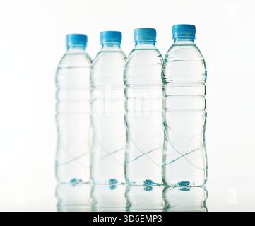Four clear plastic water bottles with blue caps stand in a straight line, casting soft reflections on the glossy surface beneath. The backdrop is a cl Stock Photo