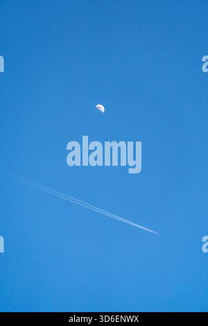 A serene image captures an airplane flying with a contrail below a bright daytime moon set against a clear blue sky, illustrating the harmony of aviat Stock Photo