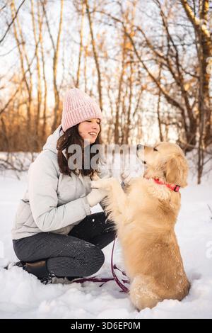 A woman wearing a beanie and winter coat kneels in the snow, joyfully engaging with a golden retriever in a snowy forest, creating a heartwarming scen Stock Photo