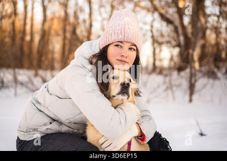 A woman in winter attire hugs her golden retriever during a snowy walk. The serene winter forest backdrop radiates warmth and companionship in the col Stock Photo