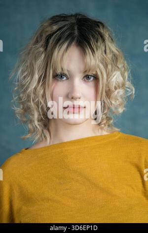 A captivating portrait of a young woman with curly blonde hair, wearing a mustard yellow sweater. The soft blue background complements her serene expr Stock Photo
