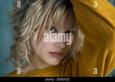 A striking portrait of a young woman with curly blonde hair and hoop earrings. The mustard yellow sweater contrasts with her cool blue grey eyes, crea Stock Photo
