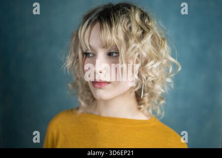 A serene portrait of a young woman with blonde curly hair, wearing a yellow top, against a soft blue background. The image captures a thoughtful expre Stock Photo