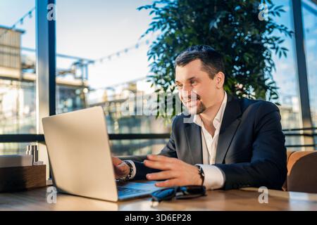 A businessman in a suit works intently on his laptop at a cafe. Sunlight streams in, creating a warm, inviting atmosphere ideal for focused work or ca Stock Photo