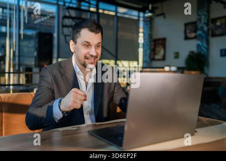 A businessman in a suit is actively working on a laptop at a cafe. He appears enthusiastic, illustrating a modern workspace outside a traditional offi Stock Photo