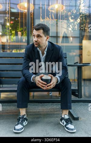 A businessman in a dark suit sits on a bench outdoors, holding a cup of coffee. He appears thoughtful and relaxed, with a modern urban setting in the Stock Photo