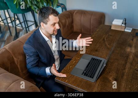 A businessman in a suit works on a laptop in a cozy cafe, engaging in a virtual meeting. The setting combines professional and relaxed environments, f Stock Photo