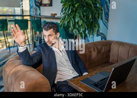 A businessman in a navy suit sits on a brown couch in a modern cafe, raising his hand as if greeting someone or calling for attention. A laptop rests Stock Photo