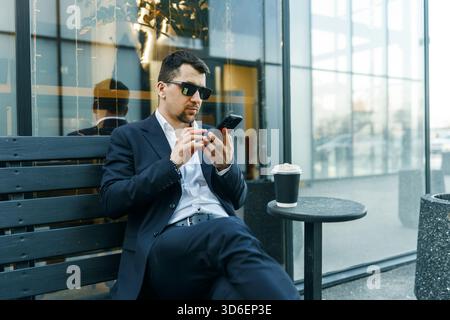 A businessman in a suit sits outdoors, using a smartphone while enjoying coffee. The modern urban setting highlights a blend of work and leisure. Sung Stock Photo