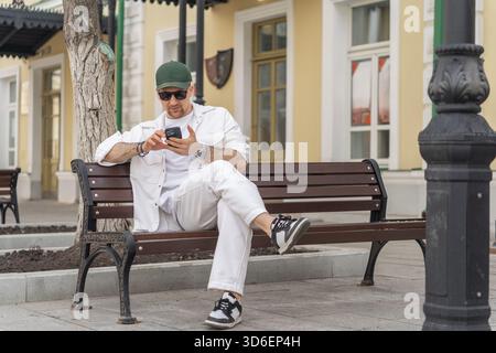 A male businessman dressed in all white attire sits on a city bench, using a smartphone, with a serene urban backdrop. Stock Photo