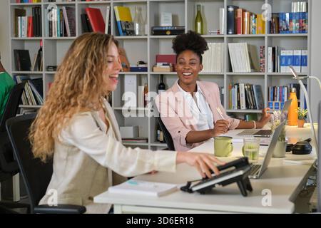 Charming diverse female coworkers sitting at table in contemporary workplace and working together Stock Photo