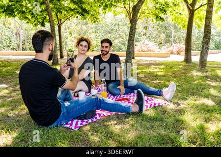 Three young adults enjoy a relaxing picnic on a vibrant blanket in the park, with one man photographing two smiling friends Stock Photo