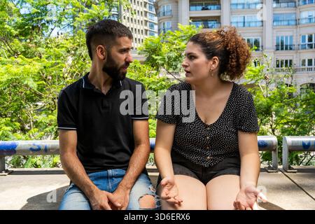 A young man and woman sit together, engaged in a serious conversation on a sunny day in an urban park, with lush greenery and modern architecture in t Stock Photo