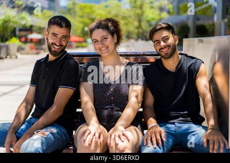 Three young adults, two men and one woman, share a joyful moment seated on a park bench under the bright sun, displaying warm smiles and casual summer Stock Photo
