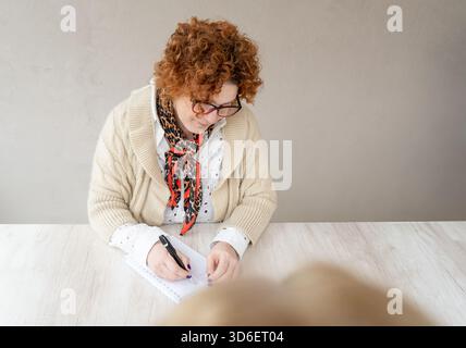A thoughtful middle-aged woman with curly hair, wearing glasses, a scarf, and a cozy sweater is captured focusing on writing notes in a spiral noteboo Stock Photo