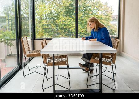 A middle-aged woman is concentrated writing notes on a modern white table, located in a bright room with a glass wall overlooking the trees Stock Photo