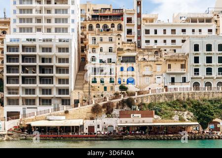 Xlendi, Gozo, Malta - April 12, 2025: Vibrant waterfront scene in Xlendi Bay with outdoor cafes, colorful jet skis and modern buildings rising behind traditional limestone houses Stock Photo