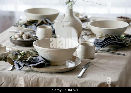 Rustic Easter table setting with empty craft ceramic tableware, plates and bowls, Easter bunny rabbit decorations, branches in vase on linen tablecloth in front of the window. Easter holiday dinner. Stock Photo