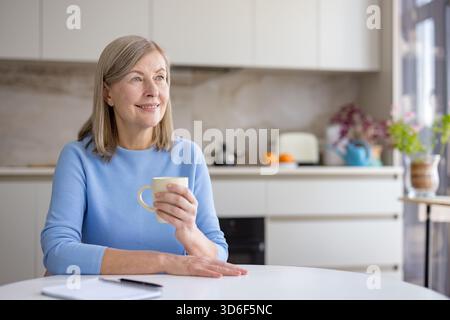 Senior woman relaxing at the kitchen table, holding a coffee mug, looking away with a thoughtful expression, enjoying a quiet moment at home during the morning Stock Photo