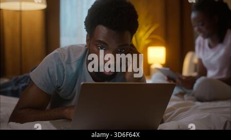 African American family couple weekend evening man boyfriend use laptop watching film lying on bed typing working woman girlfriend using digital Stock Photo