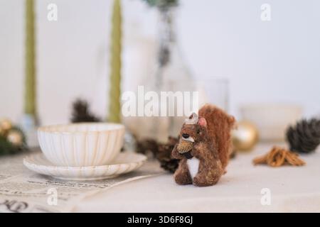 Moscow, Russia, 19.11.2025 Handmade felt squirrel holding acorn on tablecloth with pine cones and ceramic dish for autumn or winter decoration. Festiv Stock Photo