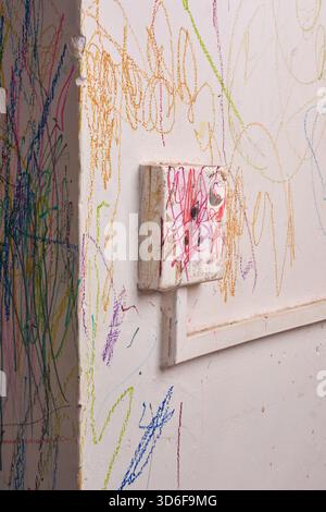 close-up of white wall covered in colorful child scribbles and worn switch box with red crayon marks, abstract messy drawings on plaster surface Stock Photo