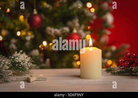 Warm Christmas candle glowing on a wooden table with festive lights and ornaments softly blurred in the background for seasonal themes Stock Photo